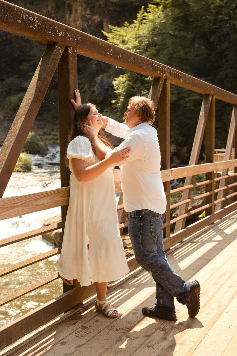 Romantic couple sharing a tender moment on a rustic wooden bridge with a river and trees in the background.