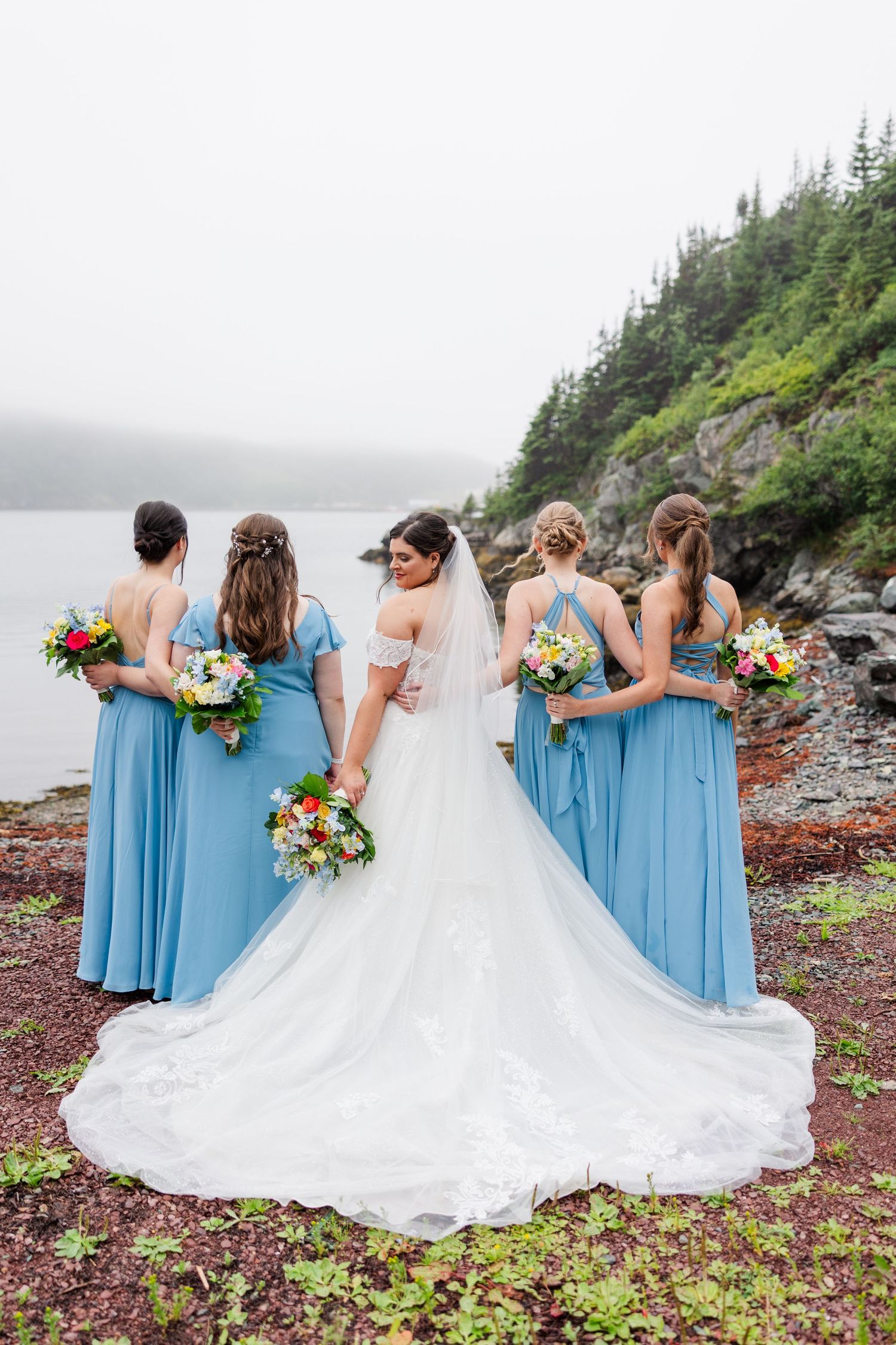Bride in white gown with bridesmaids in blue dresses holding colorful bouquets, standing by rocky shoreline with misty forest backdrop.