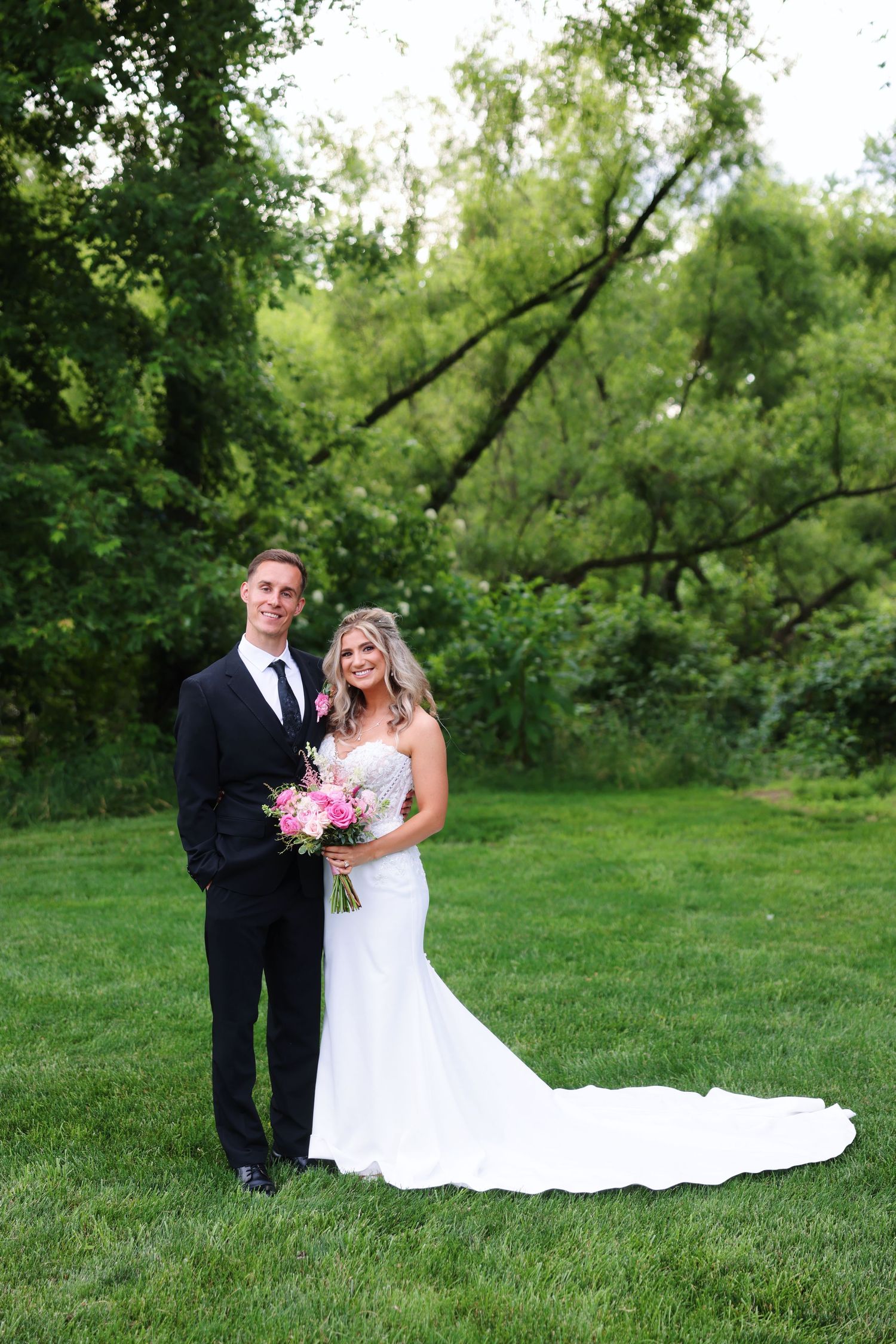Bride and groom pose together on green grass with lush trees, she holds pink bouquet in white gown with train.