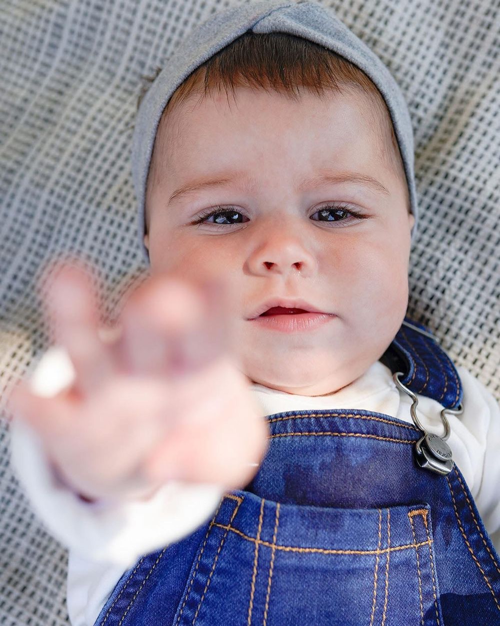 Adorable bébé en salopette en jean et chapeau gris tendant la main vers la caméra avec une expression curieuse à Pontorson.