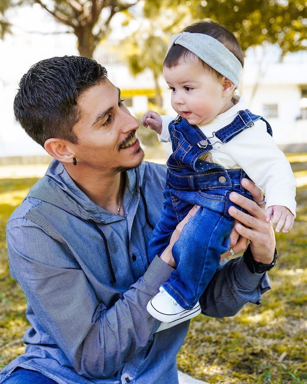 Un homme tient un bébé en salopette en jean et bandeau à l'extérieur dans un parc ensoleillé, se souriant chaleureusement à Pontorson.