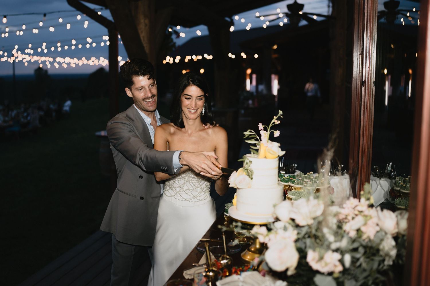 Bride and groom cutting their white wedding cake at an elegant outdoor evening reception with string lights.