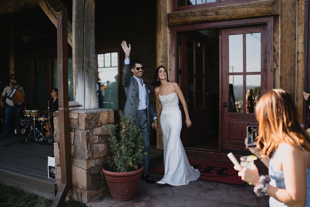 Bride and groom make grand entrance at rustic mountain wedding venue, groom waving to guests.
