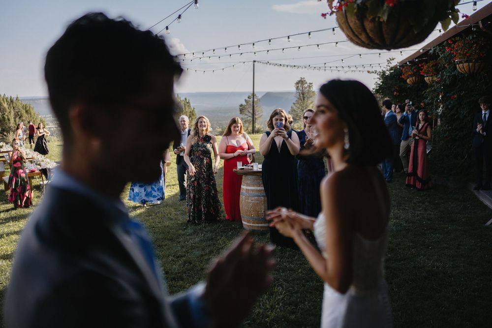 Outdoor wedding reception with guests mingling on a lawn, string lights overhead, and a lake view in the background.