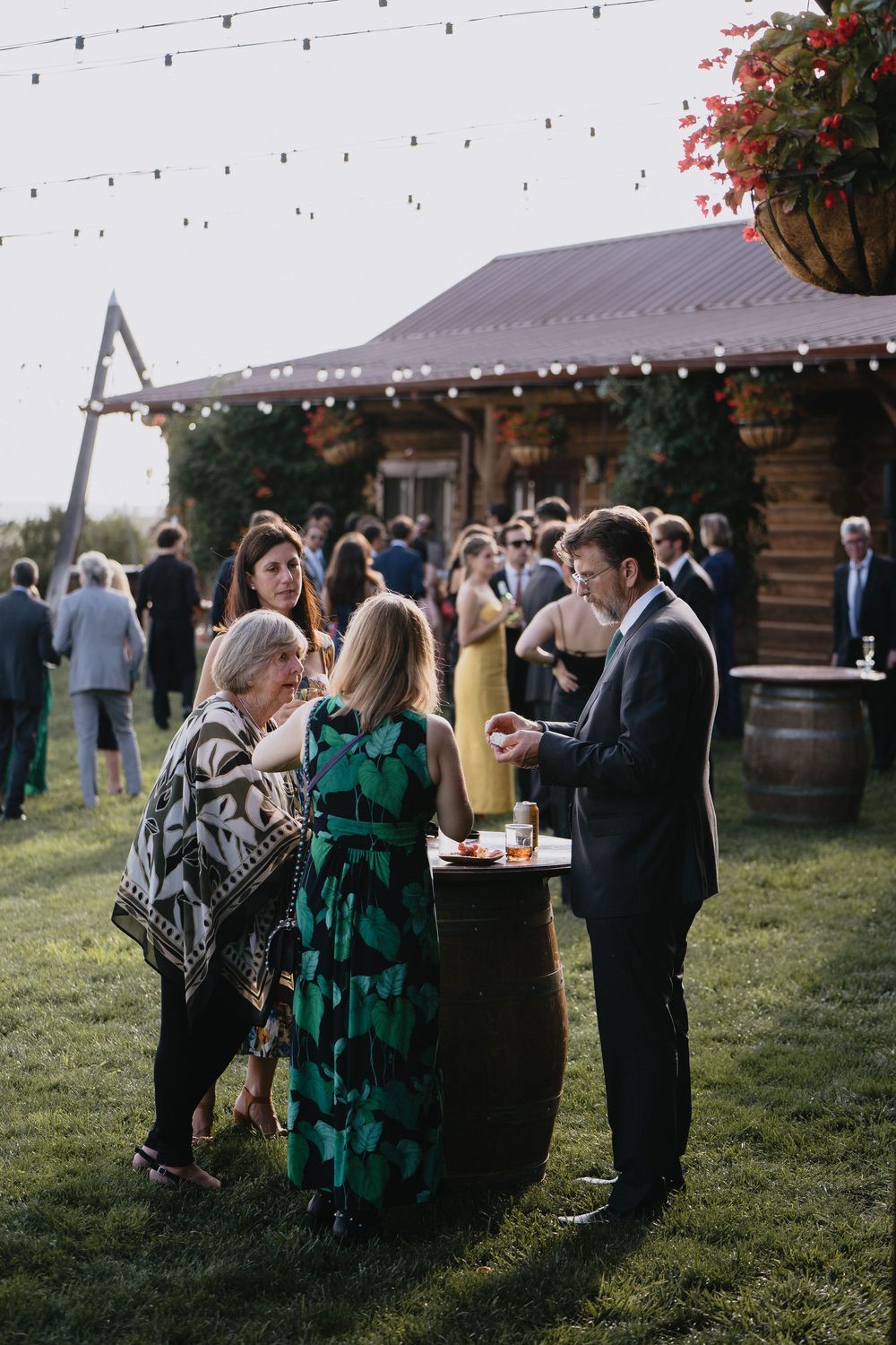 Guests mingle outdoors at a rustic barn wedding reception with string lights and wine barrel tables.