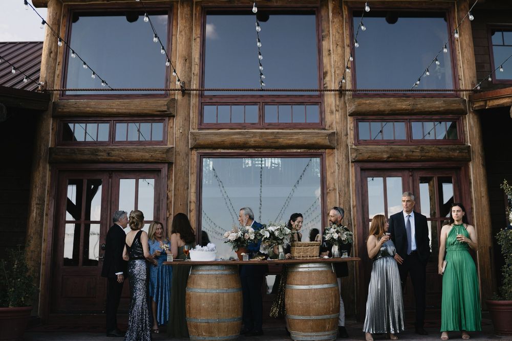 Wedding party gathered around barrel tables outside a rustic wooden venue with string lights.