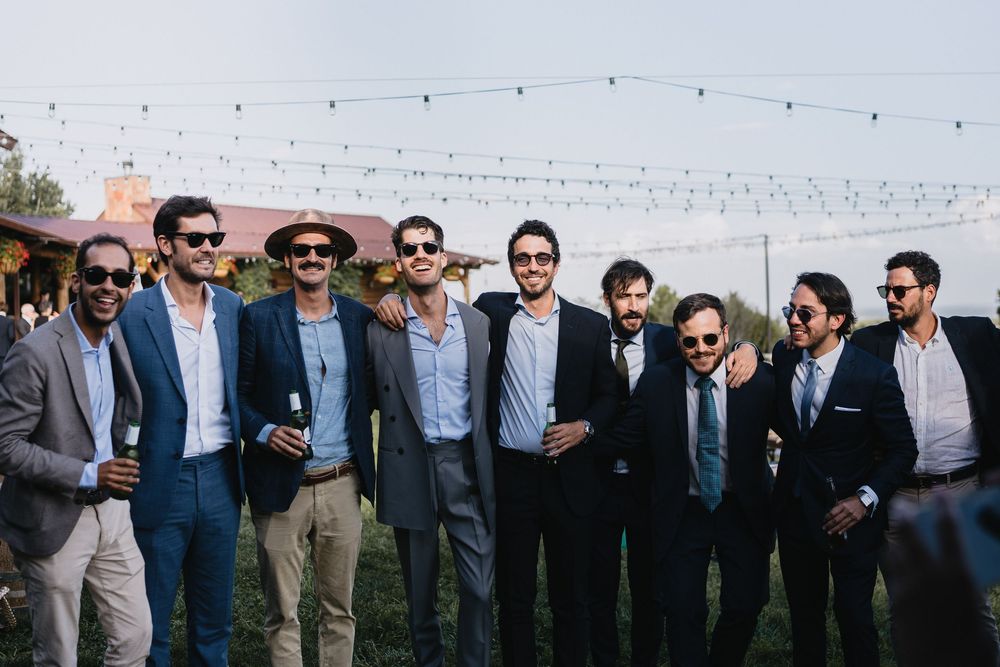 Group of groomsmen in blue suits and sunglasses smiling outdoors at a wedding reception with string lights.