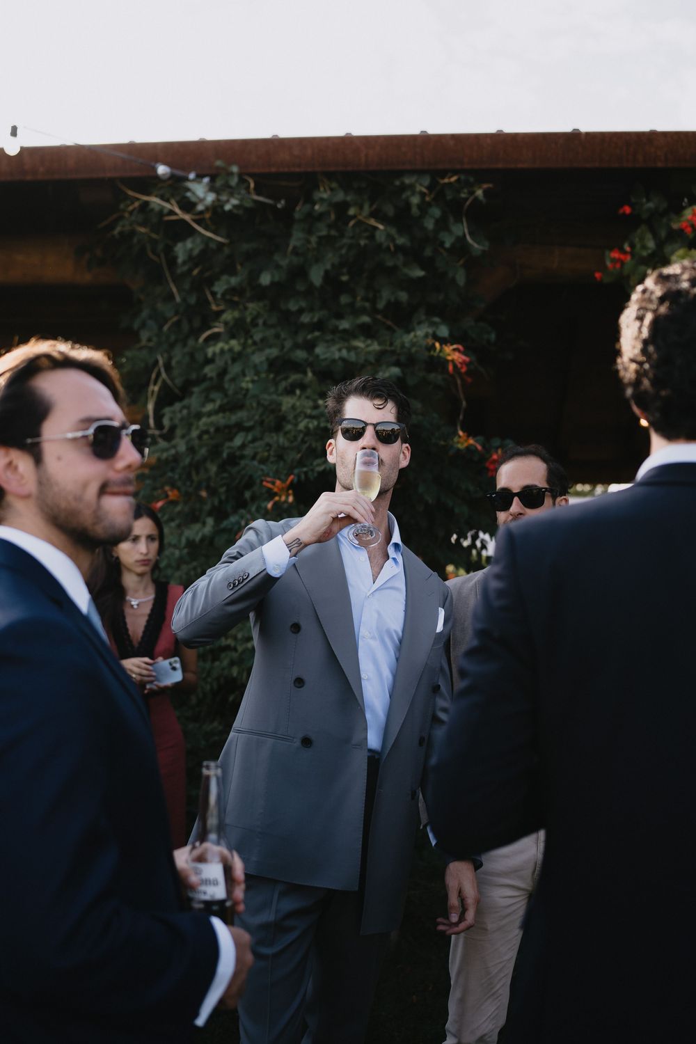 Man in gray suit raising champagne glass at outdoor wedding reception with guests in background.