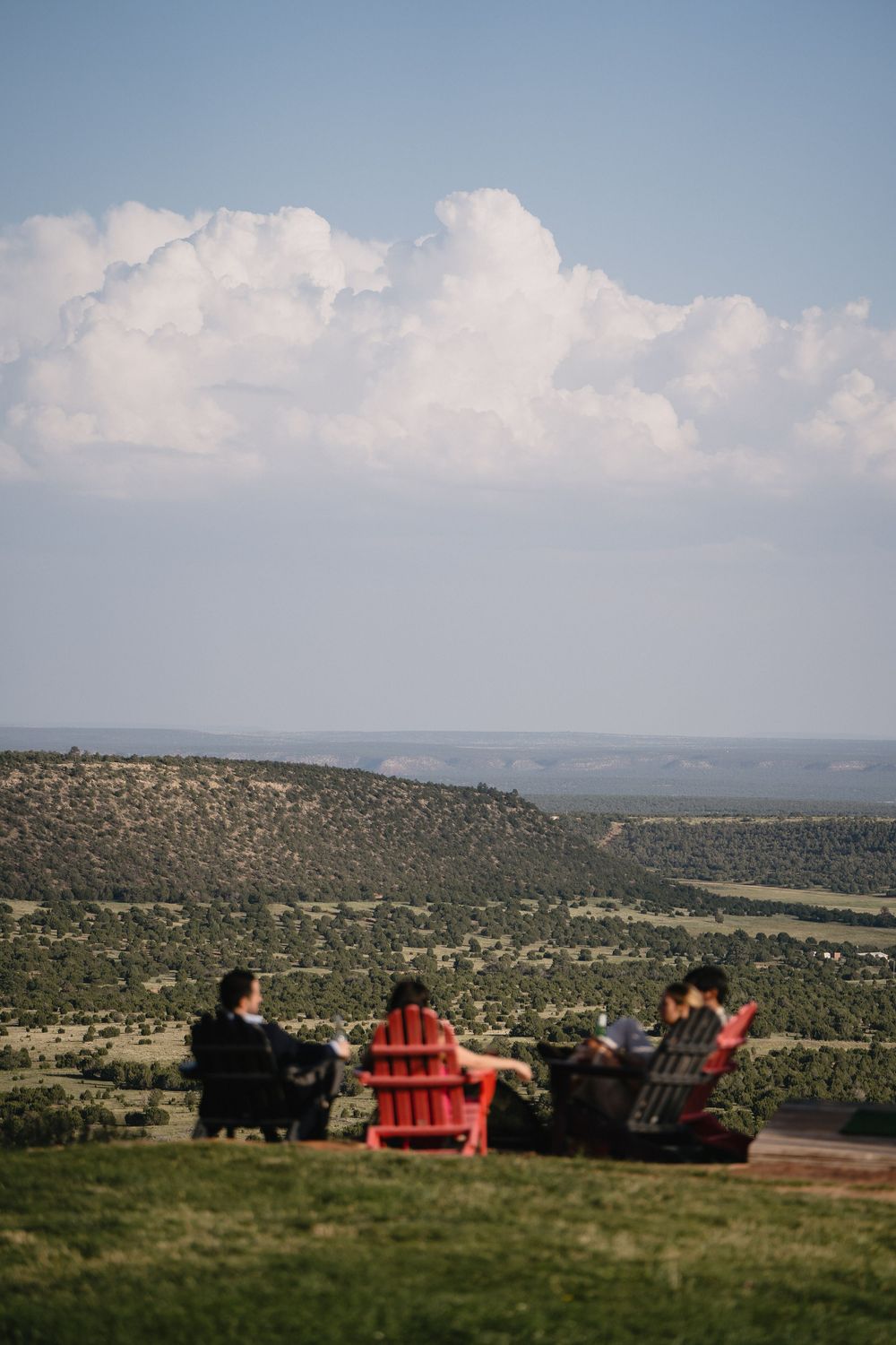Three people sitting in Adirondack chairs overlooking a vast green valley landscape under a cloudy blue sky.