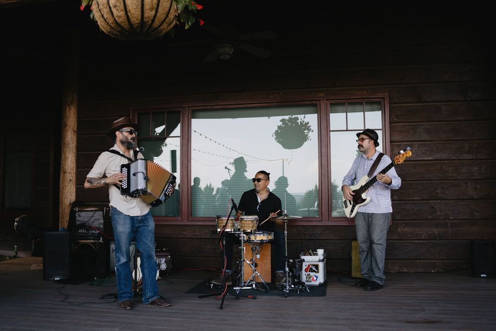 Live three-piece band playing accordion, drums, and bass guitar on a rustic wooden porch at an outdoor event.