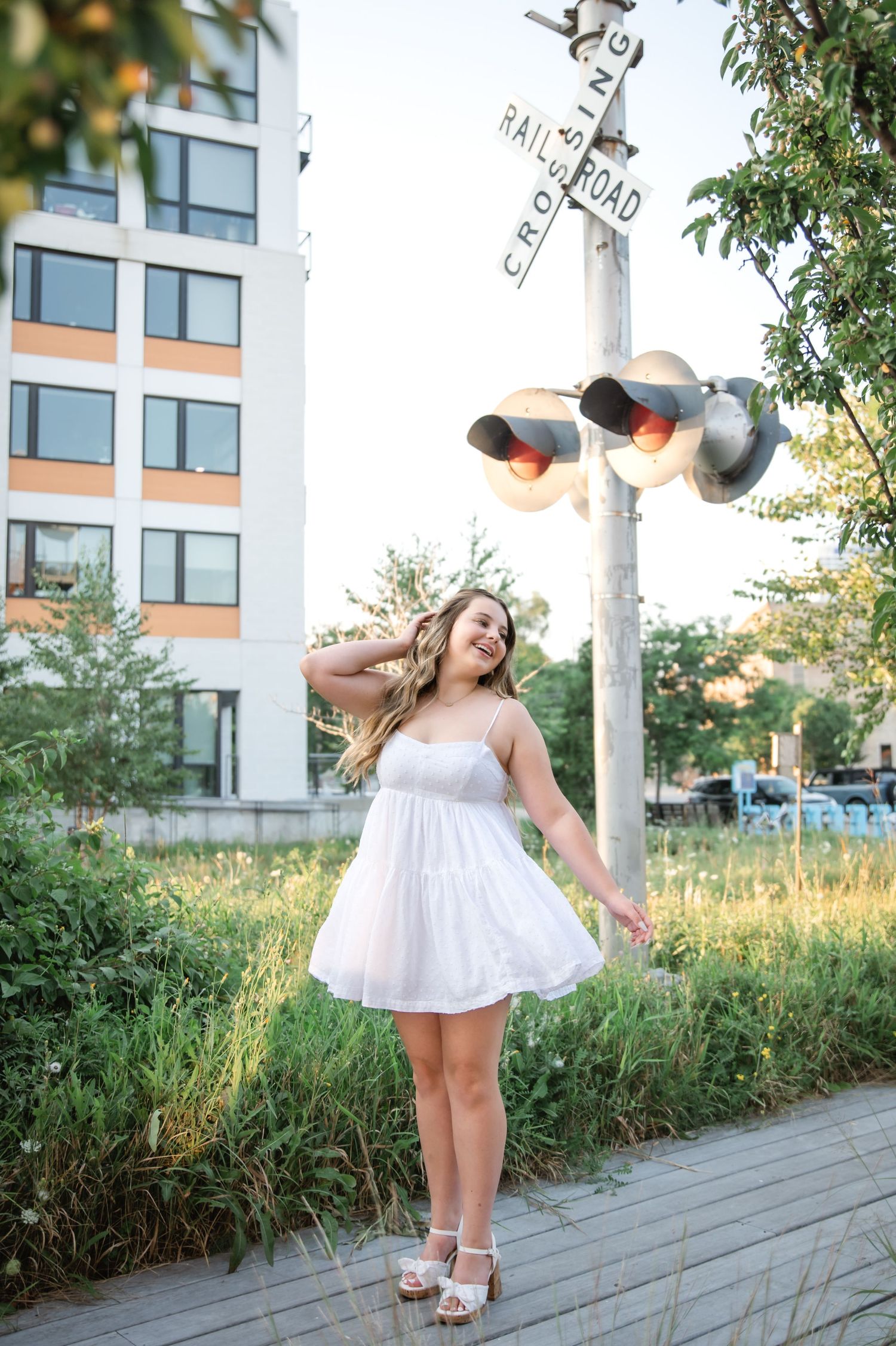Young woman in white sundress smiling near railroad crossing sign surrounded by lush greenery and urban building.