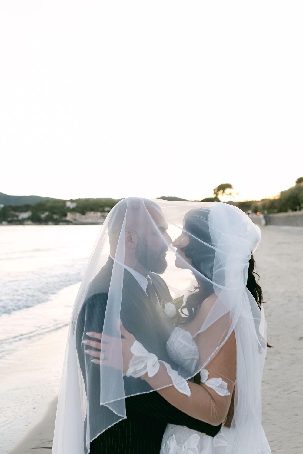 Bride and groom share a romantic kiss under a veil on a beach at sunset.
