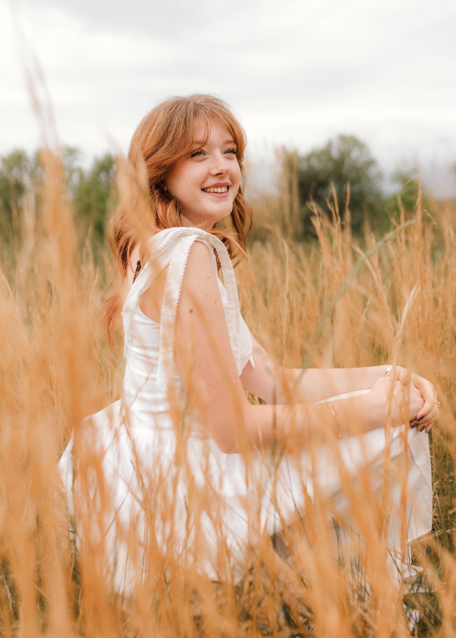 A smiling redhead woman in a white dress sits gracefully in a golden wheat field under a cloudy sky.