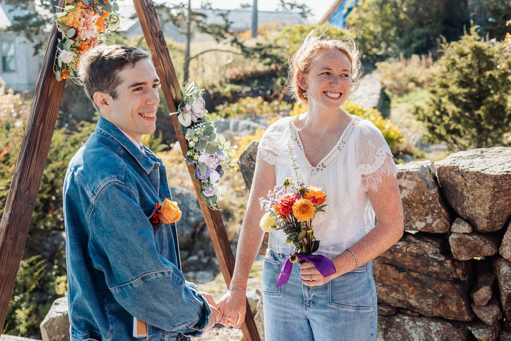 A smiling couple holds hands at an outdoor wedding ceremony with colorful floral decorations and a wooden arch backdrop.