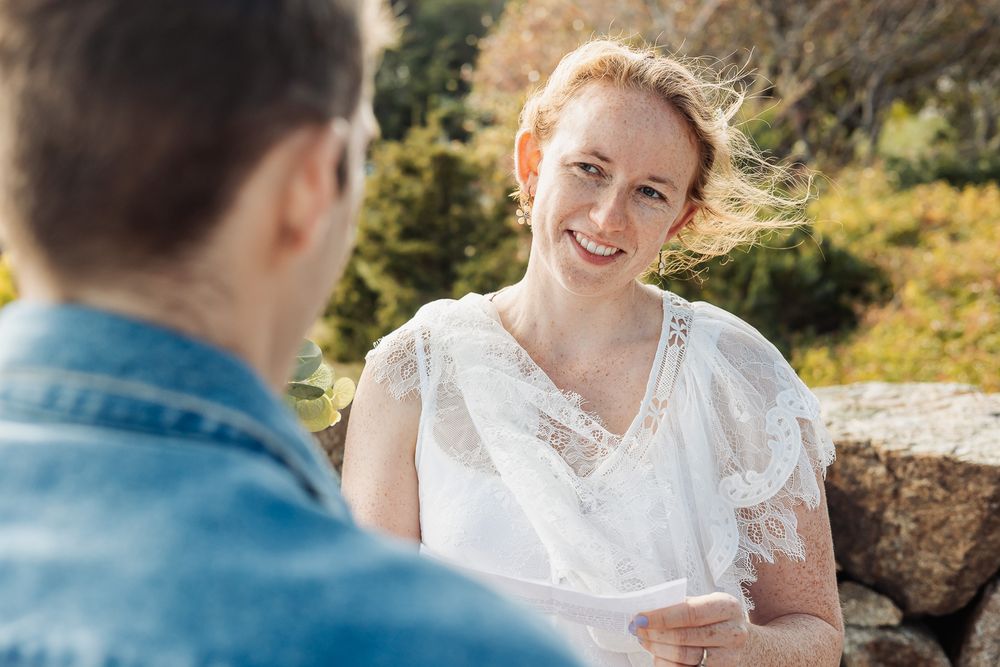 A smiling redhead woman in a white lace dress holds papers outdoors, facing a man in a denim jacket during a ceremony.
