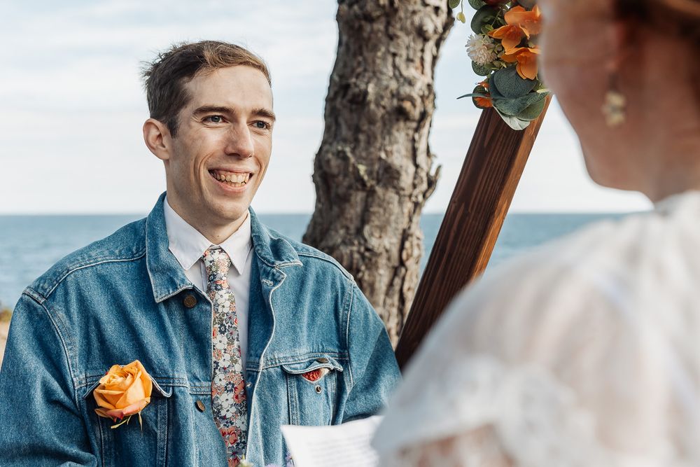 Groom in denim jacket with floral tie and orange boutonniere smiling at bride during outdoor beach wedding ceremony.