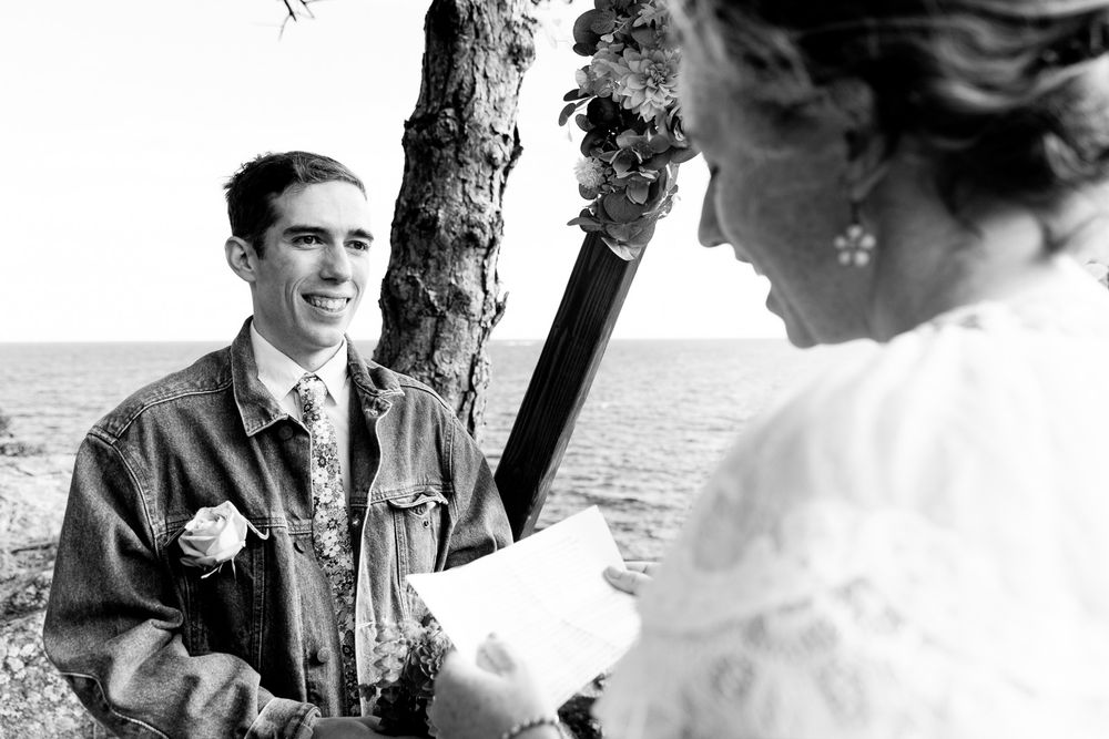 Black and white photo of a couple exchanging wedding vows outdoors near the ocean by a tree.