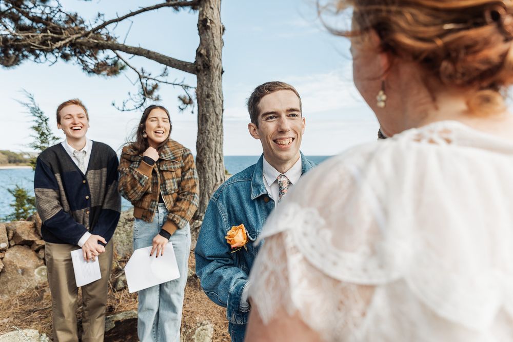 Outdoor wedding ceremony by the water with smiling guests and bride in white lace dress under pine trees.