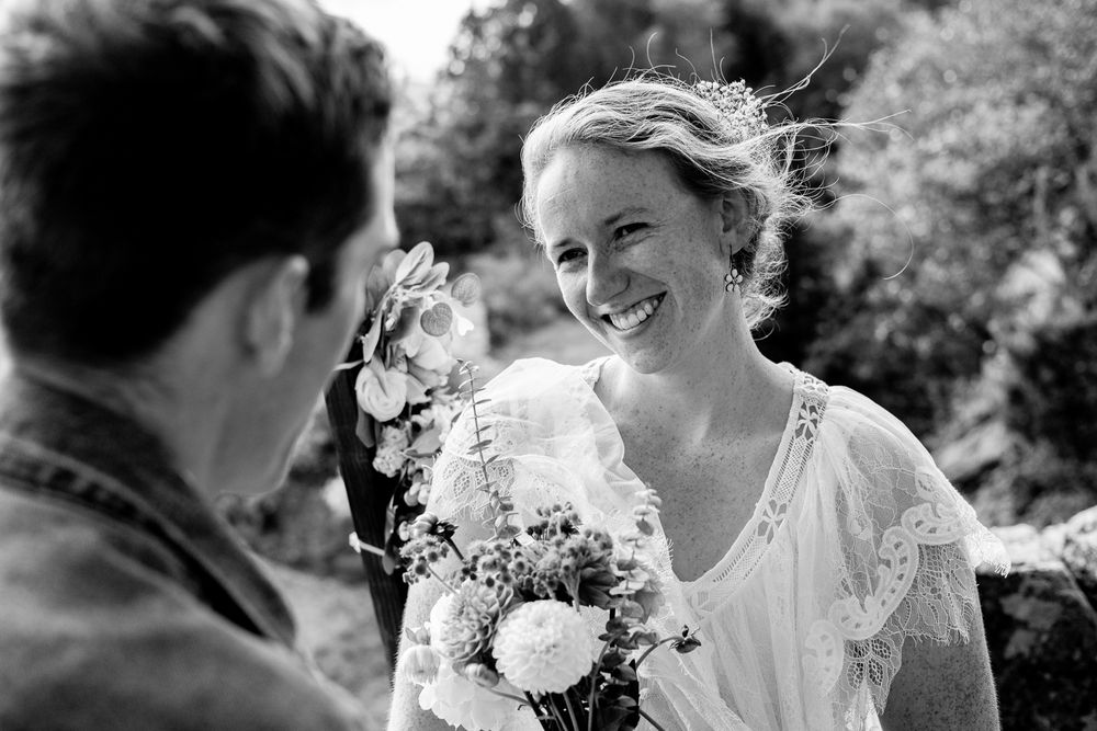 Black and white photo of a smiling bride holding a bouquet, sharing a joyful moment with her partner outdoors.