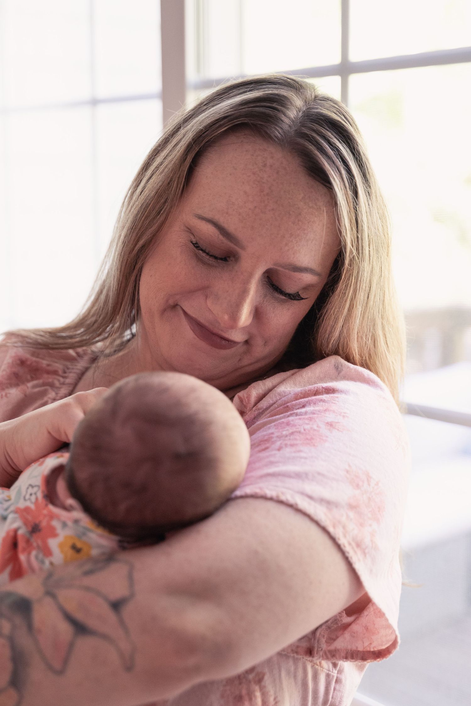 Smiling blonde mother with freckles gently cradling her newborn baby wrapped in a floral blanket.