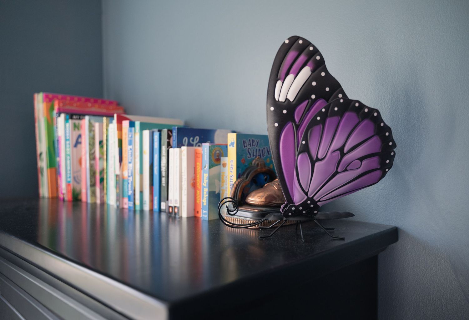 Purple butterfly decorative lamp on a dark shelf beside colorful children's books.