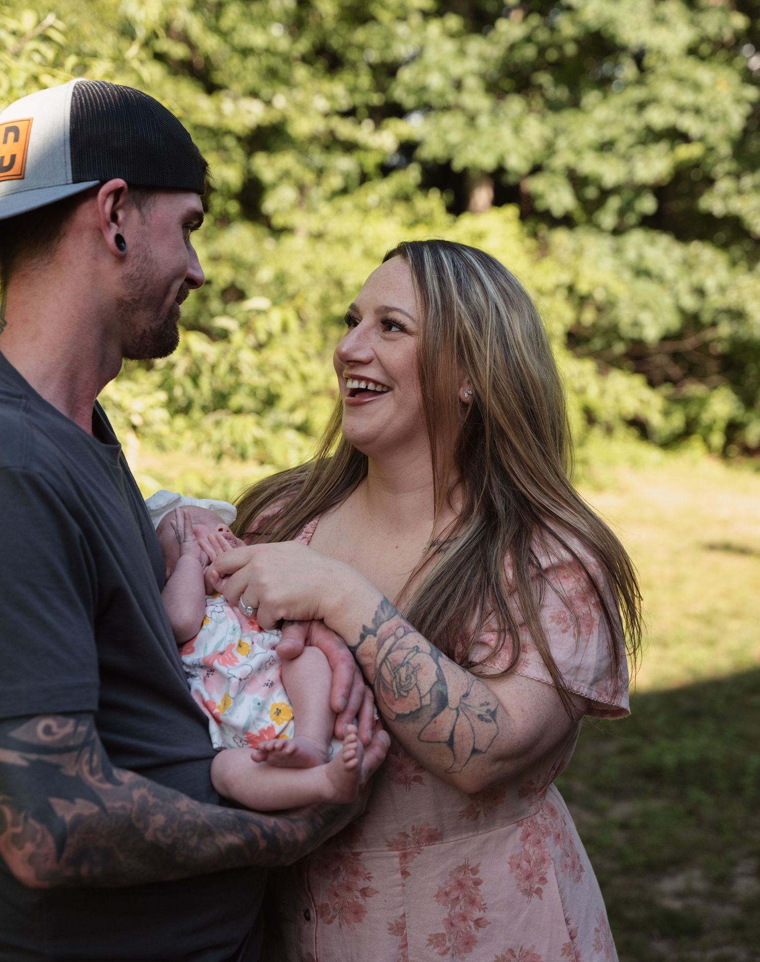 Smiling couple with tattoos holding their newborn baby outdoors in a green garden setting.
