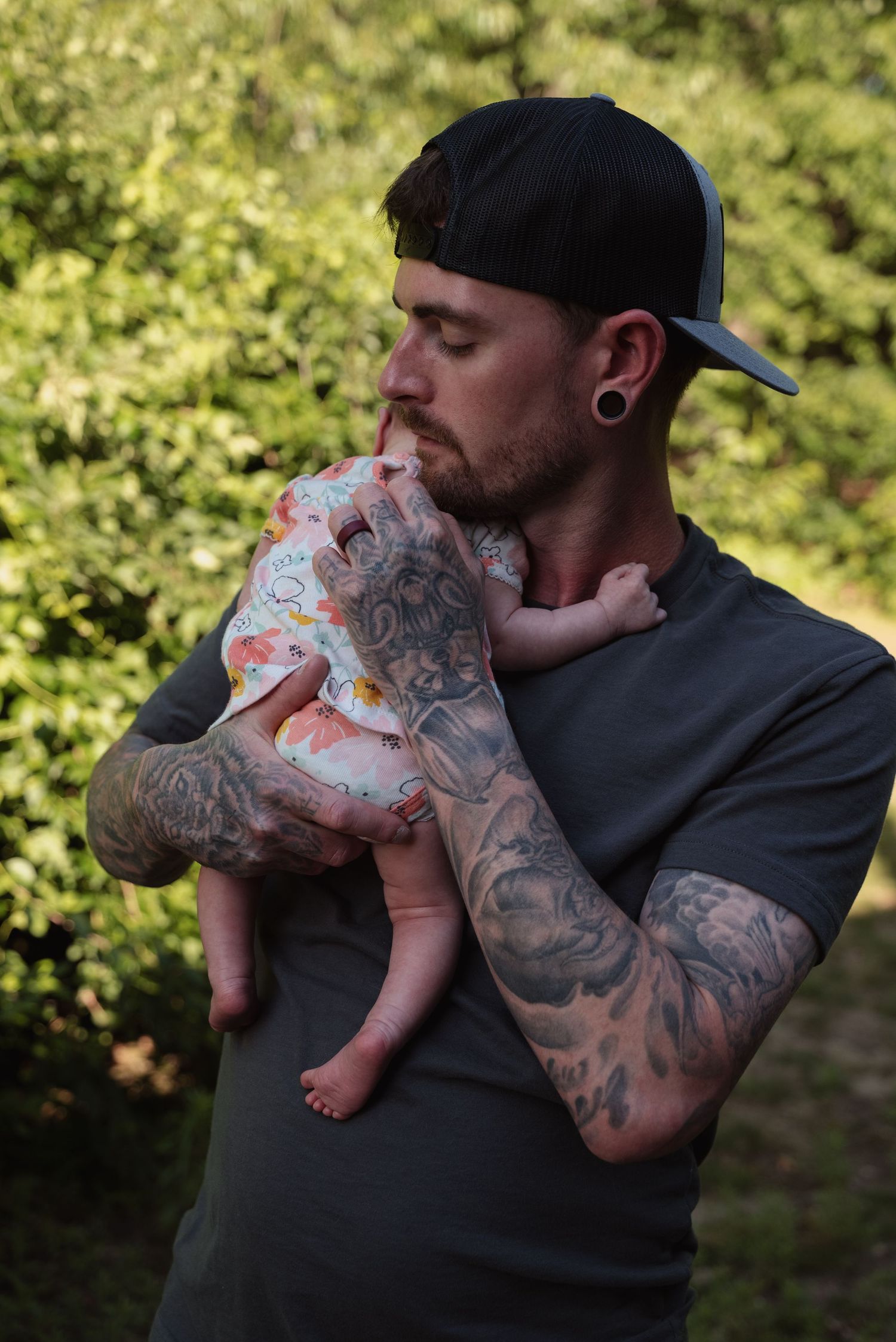 Father in gray shirt and black cap tenderly holding his newborn baby outdoors.
