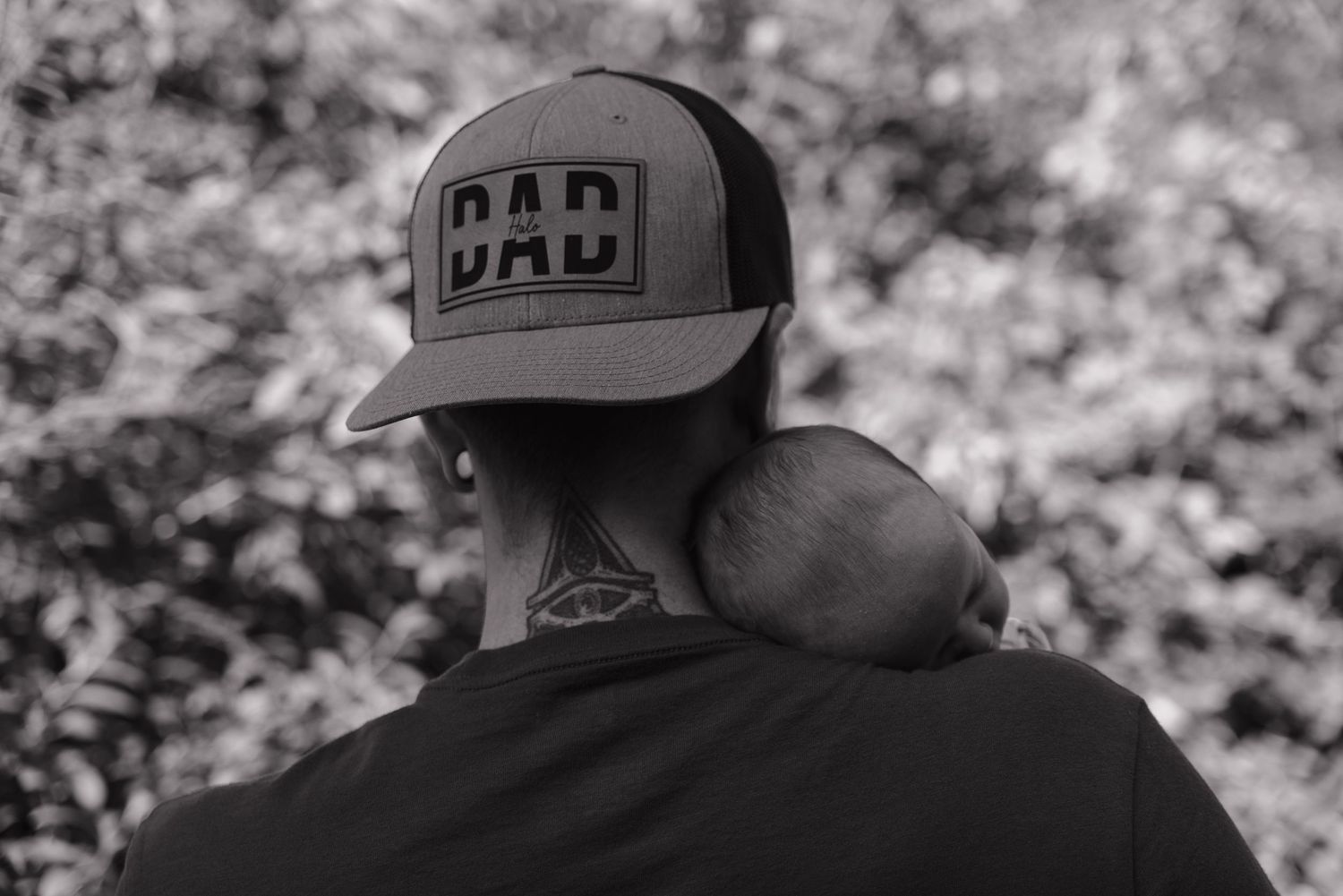 Black and white photo of a father wearing a DAD trucker hat holding a sleeping newborn.