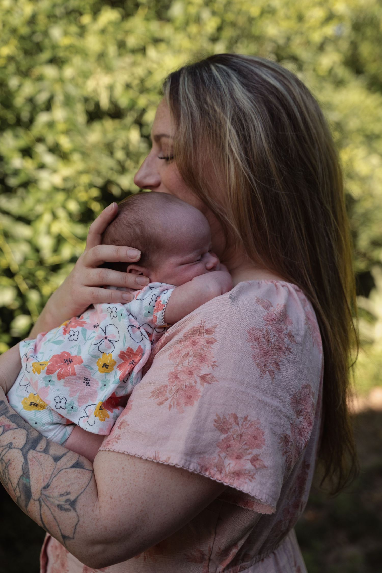 Mother in floral pink dress gently kissing her newborn baby outdoors among green foliage.