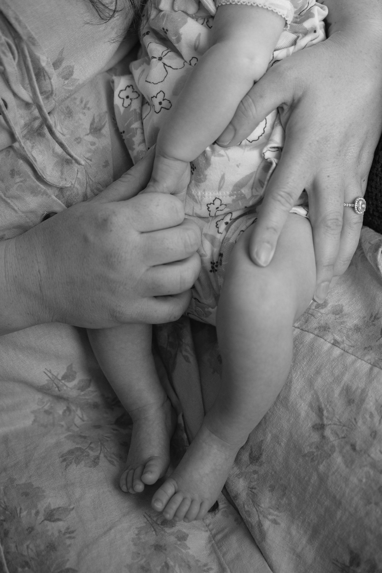 Black and white close-up of parents' hands and a newborn baby's feet on a floral blanket.