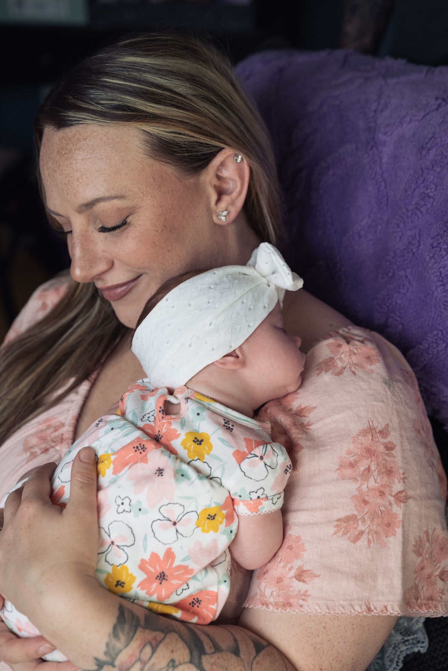 A smiling mother with freckles tenderly holds her newborn baby wrapped in a floral swaddle with a white bow hat.
