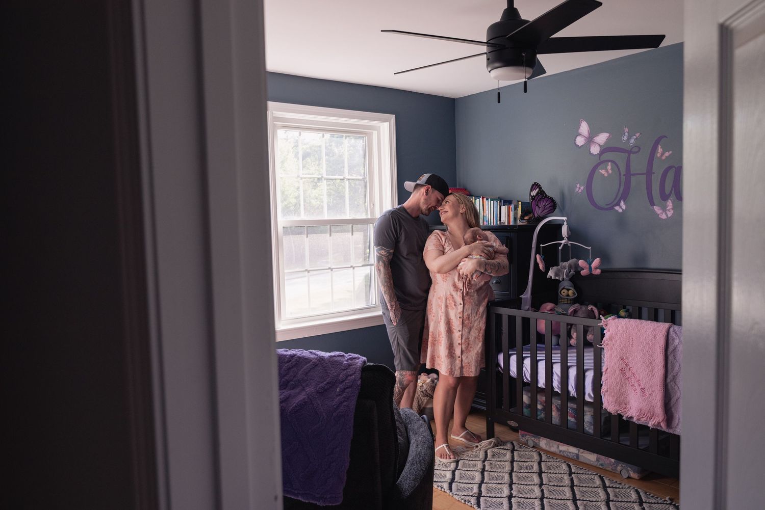 A couple shares a tender moment in a decorated nursery with a gray crib and purple butterfly wall decor.