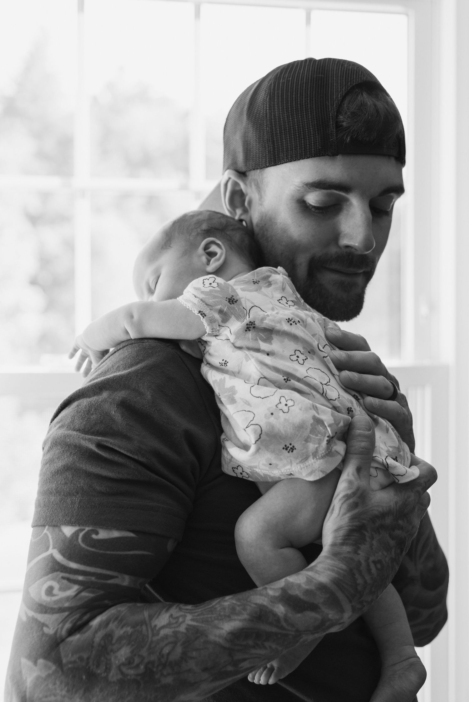 A father wearing a black cap gently holds his newborn baby against his shoulder in black and white.