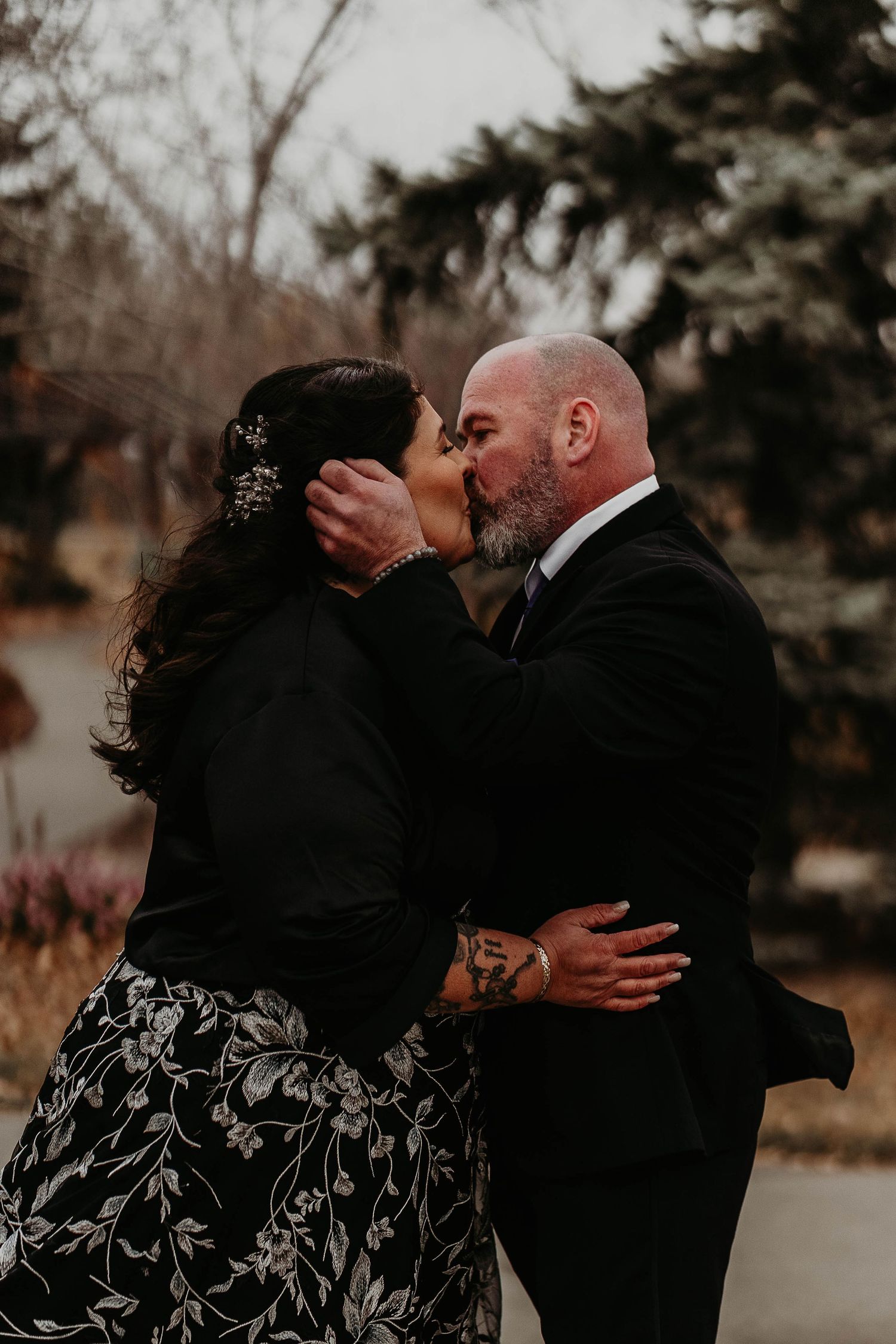 Couple embracing outdoors in winter, bride in black floral gown holding pink bouquet, groom in black suit near rustic wooden structure.
