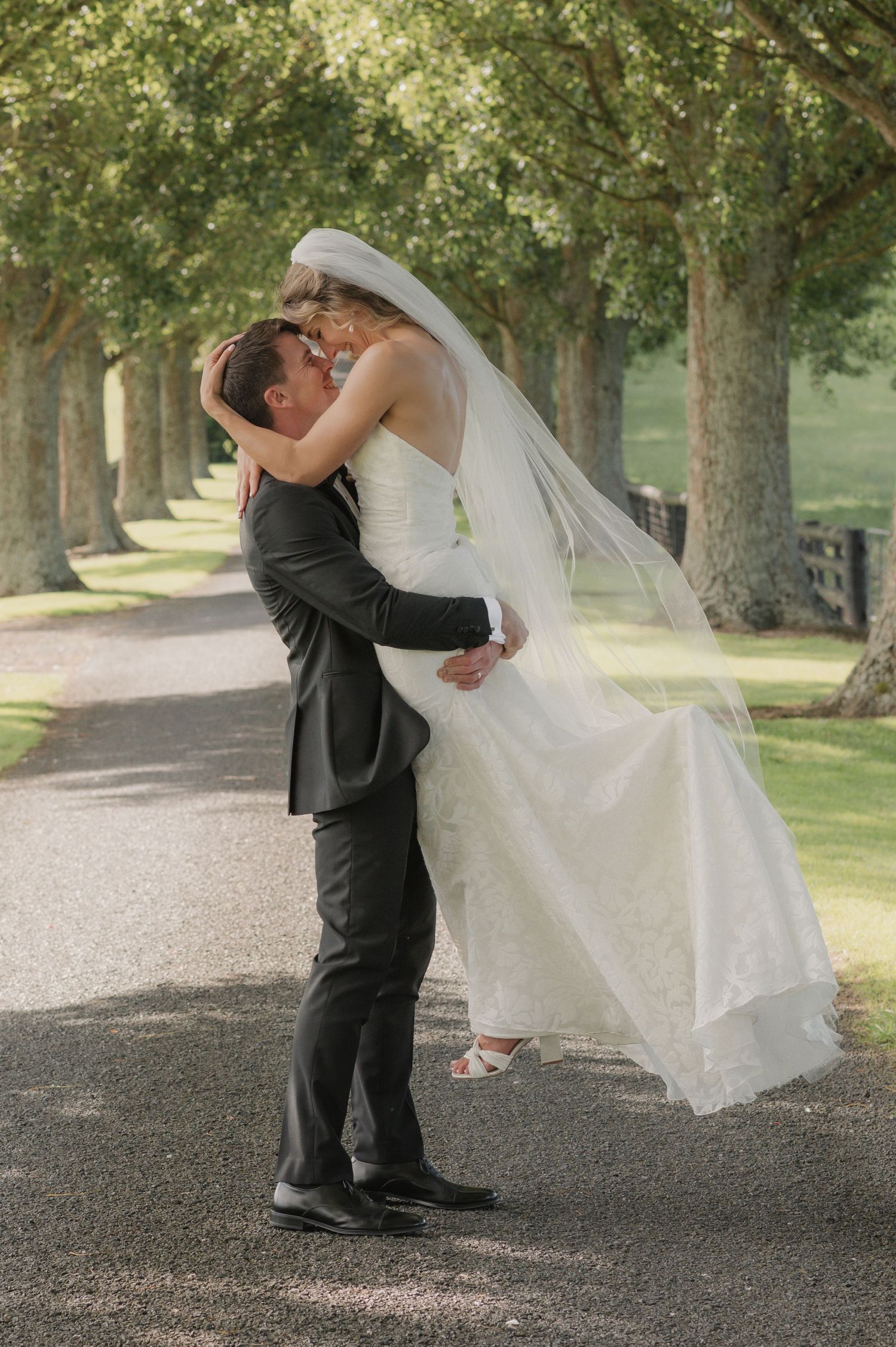 Groom lifting bride in white gown and veil, sharing a romantic kiss on a tree-lined driveway.