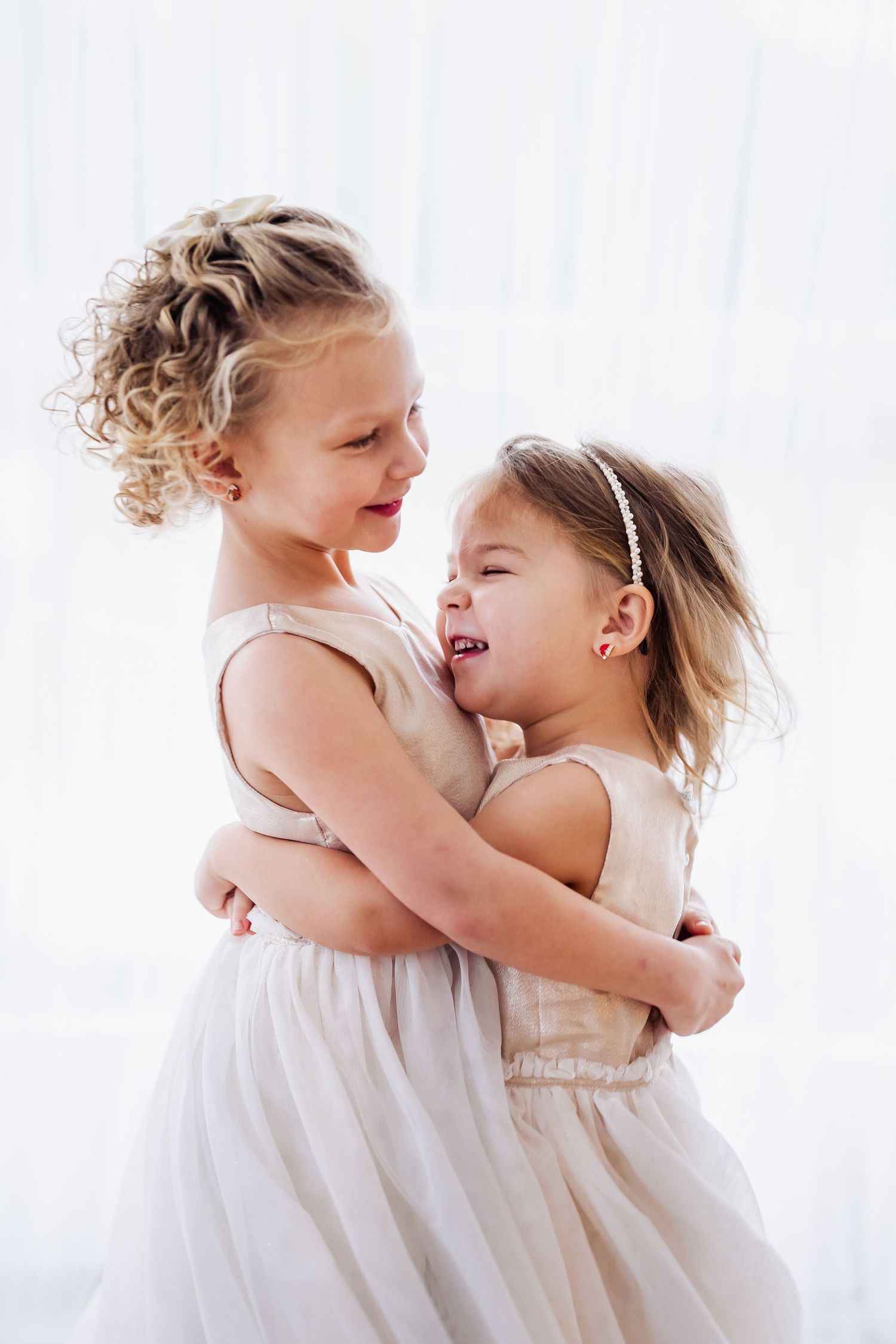 Two young girls in matching white dresses hugging and laughing together in a bright, airy room.
