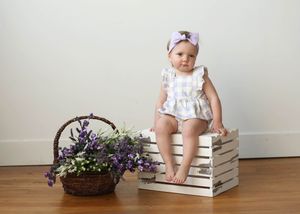 Baby girl in white romper and purple bow headband sits on white crate beside basket of purple wildflowers.