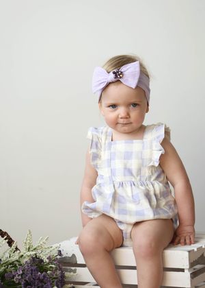 Baby girl in lavender gingham romper and purple bow headband sitting on white wooden crate.