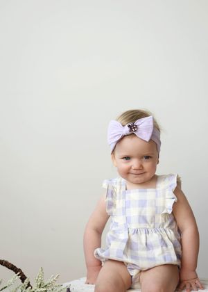 Smiling baby girl in lavender gingham romper and bow headband sitting against white background.