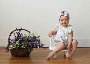 Baby girl in floral romper and pink bow headband sits on white stool beside purple flower basket.