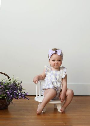 Baby girl in lavender gingham romper and bow headband sitting on white stool beside purple flowers.