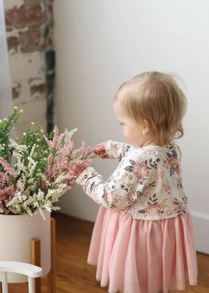Toddler girl in floral top and pink tutu dress touching white and pink flowers in a pot indoors.