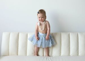 Adorable baby girl in blue tutu skirt and pearl necklace standing on white sofa.