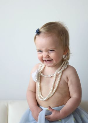 Smiling baby girl wearing pearl necklaces and a blue tutu skirt with a navy bow hair clip.