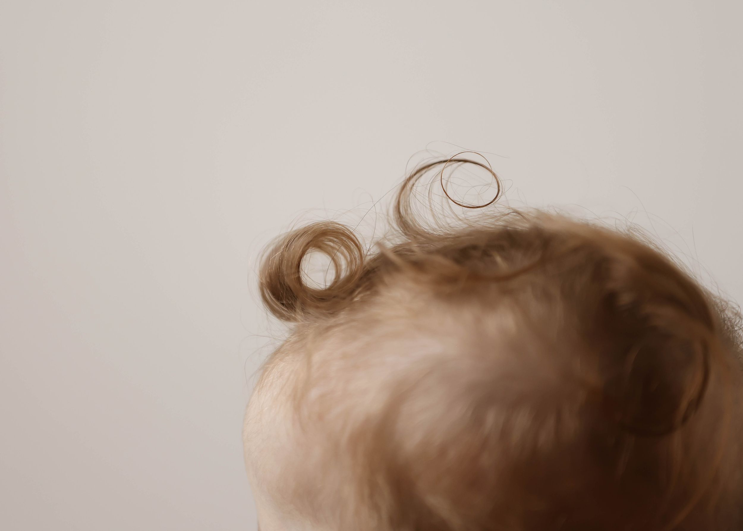 Close-up of a baby's head with soft blonde curly hair against a light background.