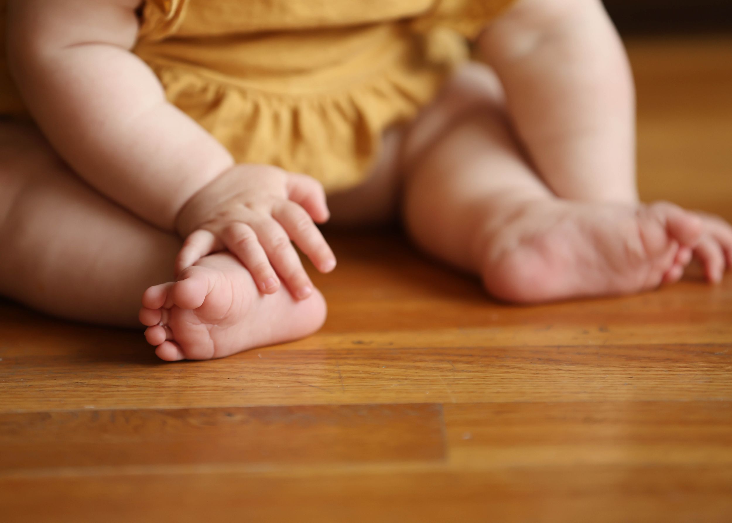 Close-up of a baby's chubby hands and feet on a warm hardwood floor, wearing a yellow dress.