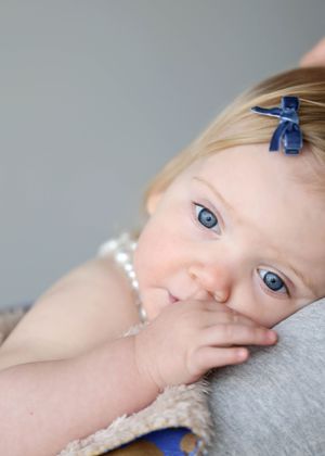 Adorable blue-eyed baby girl with a navy bow in blonde hair, resting her chin on her hand.