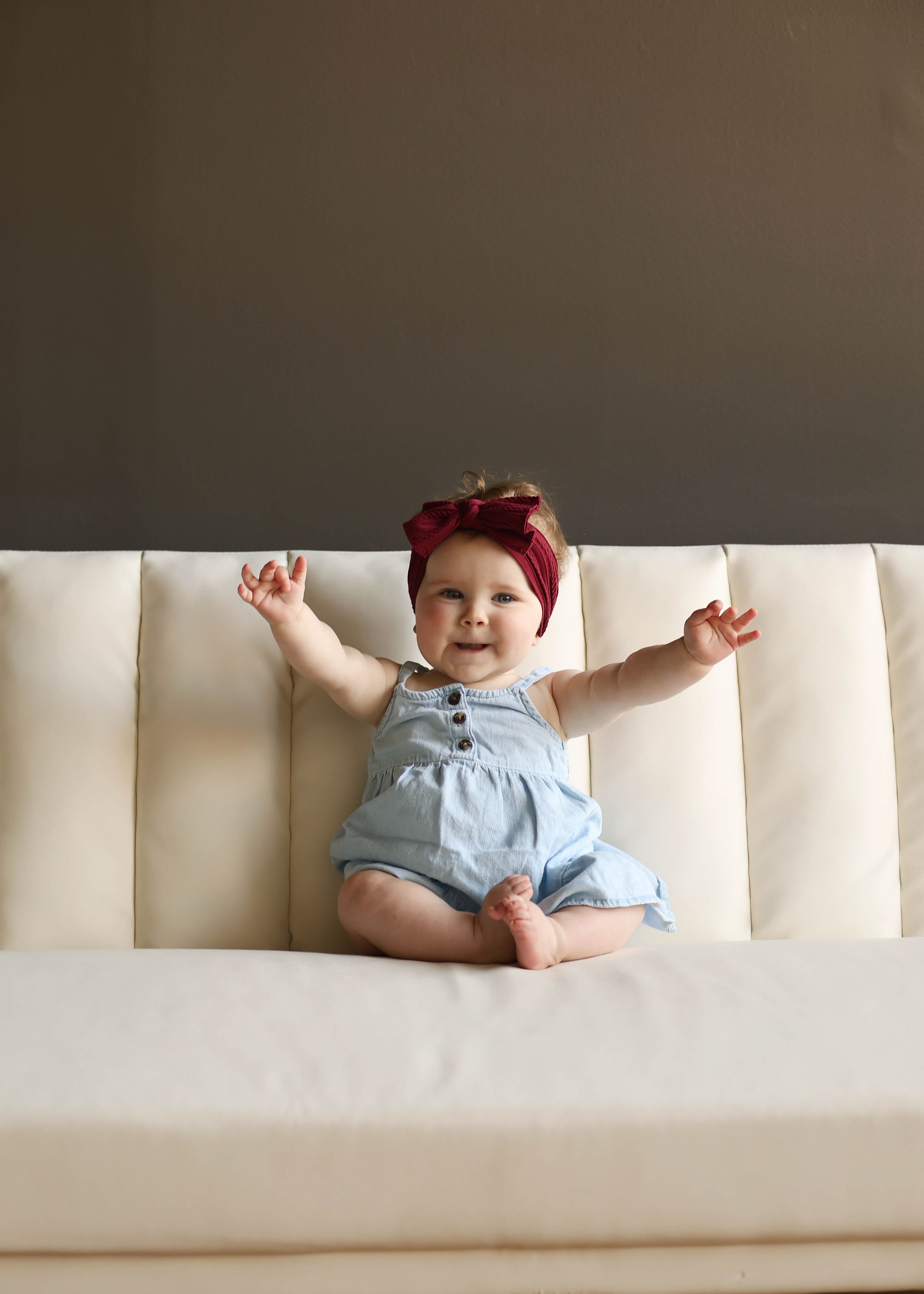 A happy baby girl in a blue romper and red bow headband sits on a cream sofa with arms raised.