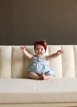A happy baby girl in a blue romper and red bow headband sits on a cream sofa with arms raised.