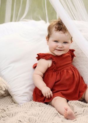 Smiling baby girl in red ruffled dress sitting on cream knit blanket with white pillows backdrop.